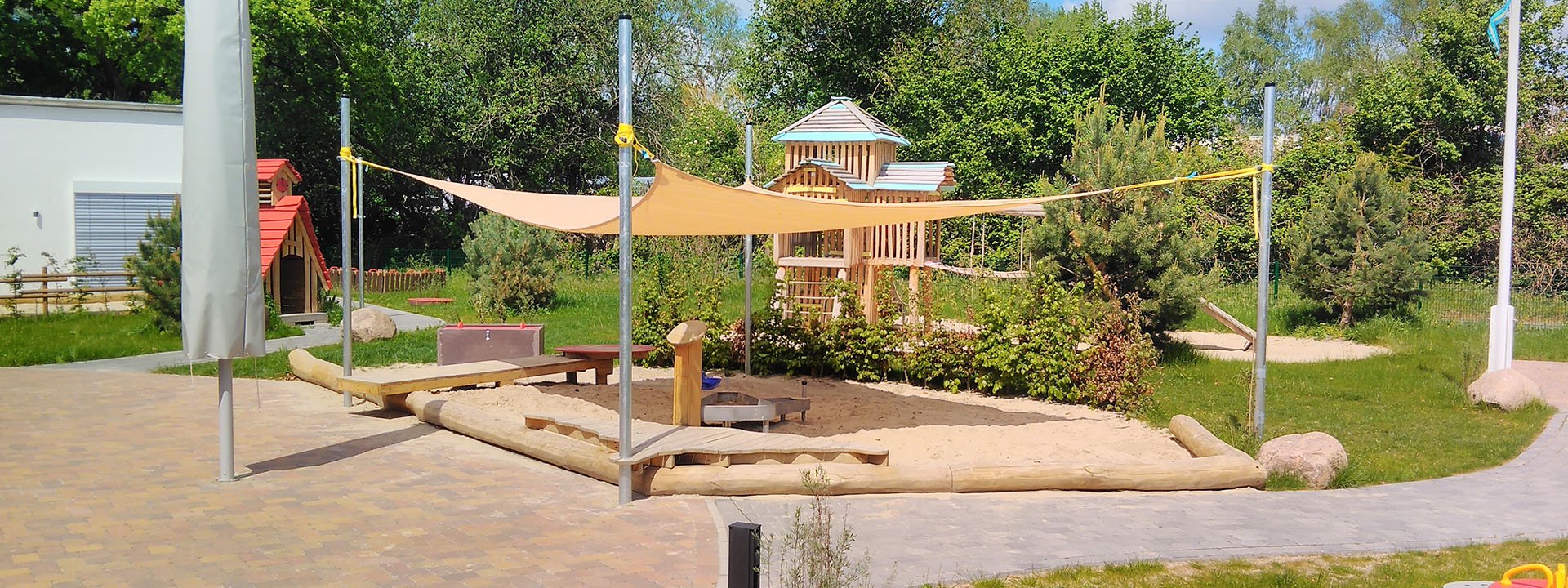 Naturnaher Sandspielplatz mit Sonnensegel, Holzspielhaus und Kletterelementen im Kindergarten