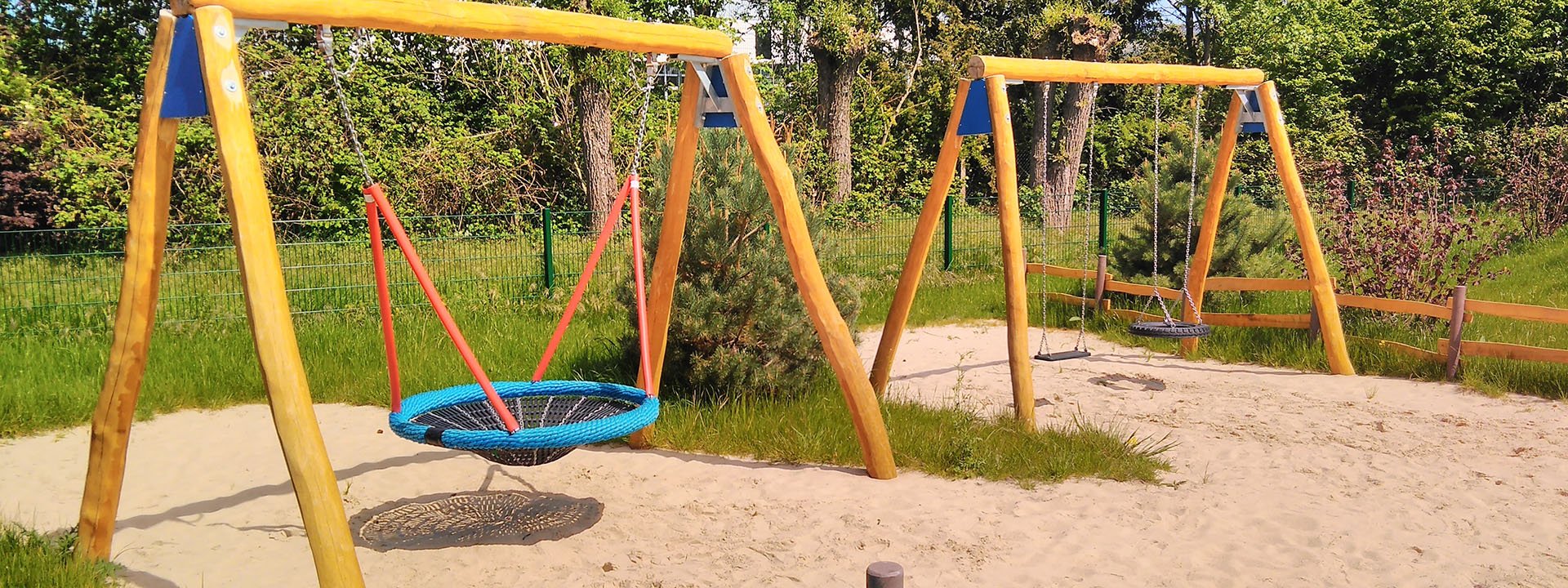 Holzschaukel mit Nestschaukel und Reifenschaukel auf naturnahem Spielplatz im Sandbereich